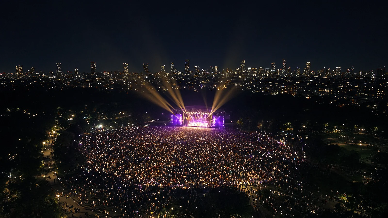 Aerial view at night of an outdoor festival stage with light beams above a vast crowd beside a city skyline
