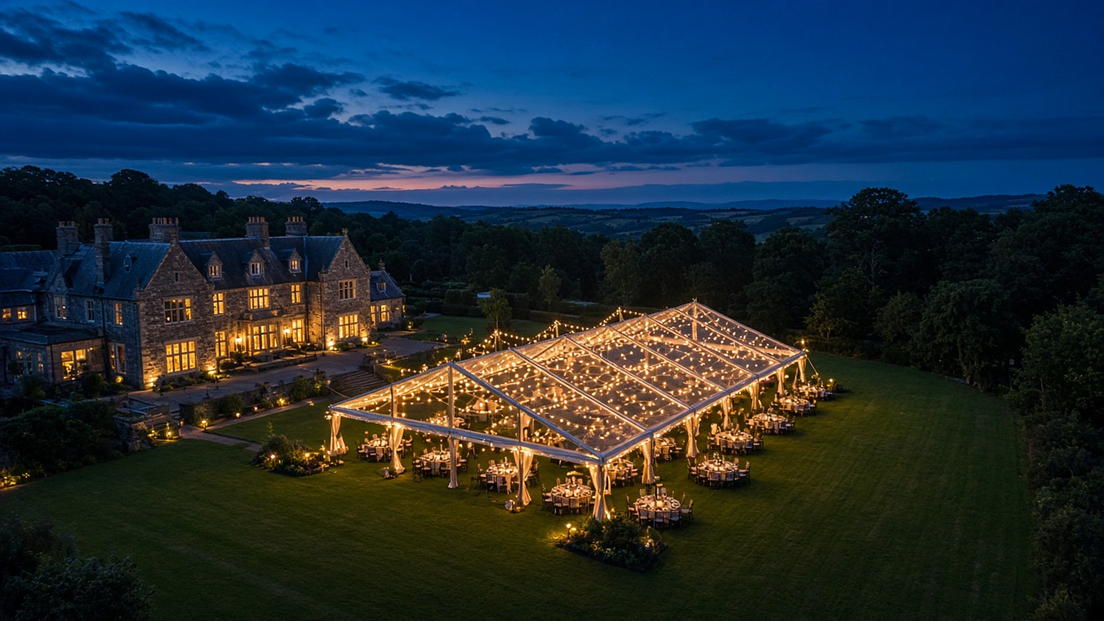 Aerial view at blue hour of a clear-top reception tent glowing with string lights beside a stone estate